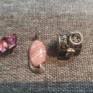 May include: Three pieces of jewelry: a faceted pink gemstone, a pink and white pendant with a silver bail, and a small silver box with ornate detailing. The items are displayed on a gray textured surface.
