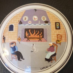 May include: A round ceramic plate with a painted scene of a fireplace with two people sitting in chairs. The plate has a brown border and the text "LAPLAU 5" in the bottom center.