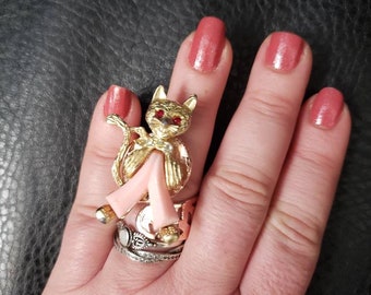 May include: A gold-colored cat ring with red gem eyes and pink legs. The cat is sitting on a stack of rings, including a silver band with clear stones. The hand has pink nail polish.