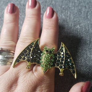 May include: A gold-toned bat ring with green and black rhinestones. The bat's wings are adorned with black stones, while the head features green stones. The ring is a statement piece, perfect for Halloween or adding a touch of whimsy to any outfit.