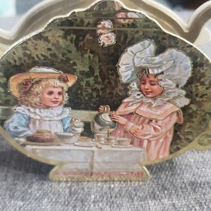 May include: A gold teapot-shaped tea bag holder with a painted scene of two girls having tea in a garden. The girls are wearing dresses and hats, and the scene is surrounded by green foliage.