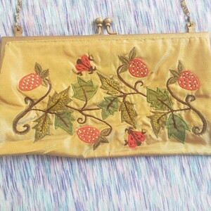 May include: A golden clutch with a chain strap, featuring an embroidered design of strawberries, leaves, and vines. The strawberries are red with white dots, and the leaves are green and brown. The clasp is a vintage style.