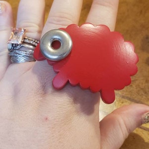 May include: A red sheep-shaped ring with a silver ring in the center. The ring is worn on a person's finger.