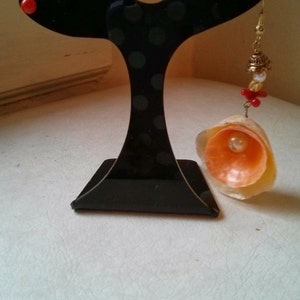 May include: A pair of earrings with a seashell pendant and a gold chain. The seashell is orange and white with a pearl in the center. The earrings are displayed on a black polka dot stand.