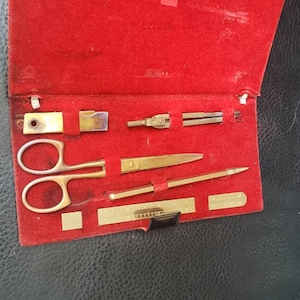 May include: A red velvet case with a set of gold-toned tools, including a pair of scissors, a compass, a ruler, and a small knife. The tools are arranged neatly in the case.