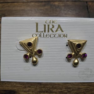 May include: A pair of gold-tone earrings with purple stones. The earrings are shaped like a triangle with a drop-shaped pendant. The earrings are on a white card with the text "The Lira Collection".