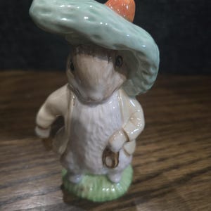 May include: A ceramic figurine of a rabbit wearing a light green hat with an orange accent. The rabbit is dressed in a cream-colored jacket and stands on a patch of green grass. The figurine has a detailed, hand-painted appearance.