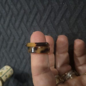May include: Two stacked rings featuring a smooth, polished finish with a brown and tan tortoiseshell pattern. The top ring has a flat, rectangular profile, while the bottom ring is rounded. Both rings are worn on a finger, showcasing the pattern.