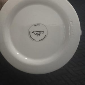May include: A white ceramic mug base with a circular logo featuring a bird design. The logo is surrounded by text that reads "HAND WASH" and "MADE IN CHINA". The mug is made in China.