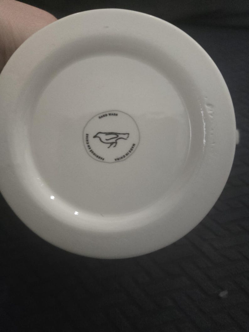 May include: A white ceramic mug base with a circular logo featuring a bird design. The logo is surrounded by text that reads "HAND WASH" and "MADE IN CHINA". The mug is made in China.