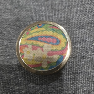 May include: A round, gold-toned compact mirror with a colorful paisley design. The fabric insert features a pattern of yellow, blue, pink, and green. The compact is set against a gray textured background.
