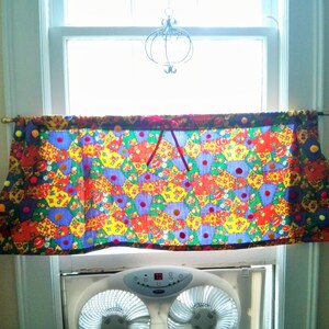 May include: A colorful window valance with a patchwork design of hexagonal shapes in red, yellow, blue, and green. The valance is trimmed with small, colorful pom-poms and hangs from a gold-colored rod.
