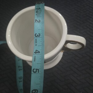 May include: A white ceramic mug with a gold handle and trim, measured with a blue measuring tape. The mug is approximately 5 inches tall. The mug is on a dark surface.