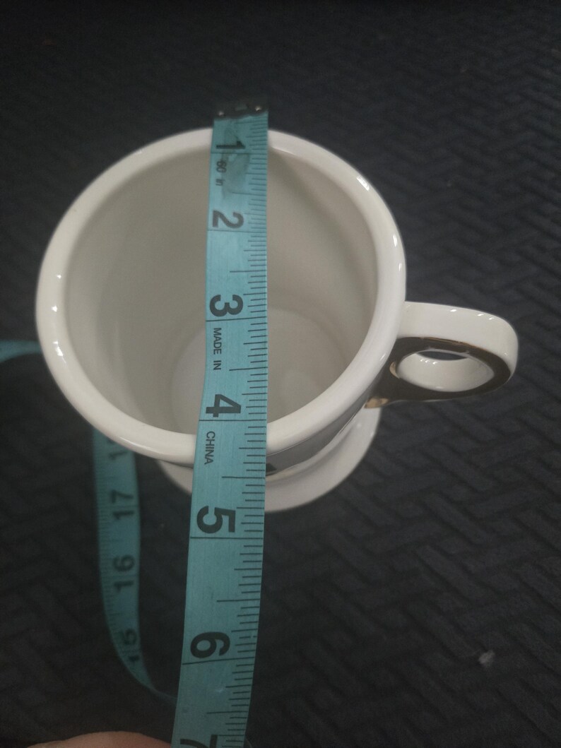May include: A white ceramic mug with a gold handle and trim, measured with a blue measuring tape. The mug is approximately 5 inches tall. The mug is on a dark surface.