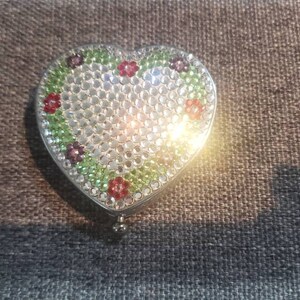 May include: A heart-shaped compact mirror with a white background and a green and red floral design. The mirror is covered in rhinestones.