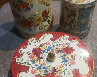 May include: Three vintage tin containers with floral designs. The containers are round and have lids. The lids are decorated with a floral pattern and have a knob on top. The containers are white with red, gold, and blue accents.