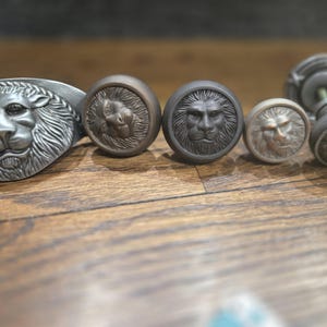 May include: A collection of antique-style cabinet hardware featuring lion head designs. The hardware includes a long pull and several round knobs in various metallic finishes, including silver, bronze, and pewter. The lion faces are intricately detailed.