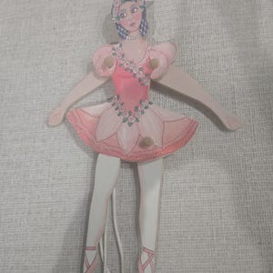 May include: A wooden ballerina doll with a pink dress and ballet shoes. The doll has a painted face, blue hair, and a floral headpiece. The arms and legs are jointed, and the doll is suspended by a string.