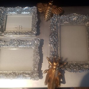 Puede incluir: Tres marcos de fotos de plata adornados con una abeja dorada y una figurita de insecto dorada sobre una superficie blanca.