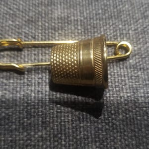 May include: A gold-colored thimble attached to a safety pin. The thimble has a textured surface and a smooth band around the top.