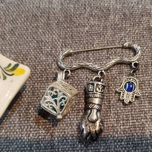 May include: A silver safety pin with five charms: a small cage, a hand with an eye, a hand with a blue eye, a small fan, and a hand with a ring. The charms are hanging from the pin.