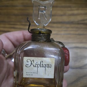 May include: Vintage perfume bottle with a clear glass stopper featuring the letter "R". The bottle has a label that reads "Réplique, Raphael, Paris". The bottle is a light amber color and has a decorative cord around the neck.
