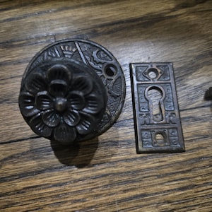 May include: Antique bronze door hardware set, including a floral doorknob, a decorative backplate, and a keyhole cover. The set is made of metal and has a dark, aged finish. A screw is also included. The hardware is set on a wooden surface.