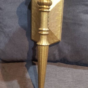 May include: A gold-toned metal wall sconce with a textured finish. The sconce has a long, slender arm and a decorative, rounded top.