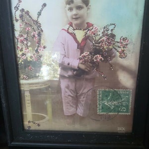May include: A framed vintage photograph of a young boy in a pink shirt holding a bouquet of flowers. The photo is decorated with glitter and has a postage stamp in the corner.