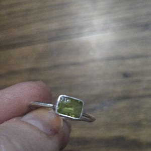 May include: A silver ring featuring a rectangular, light green gemstone. The gemstone is set in a square bezel. The ring is held against a wooden background.