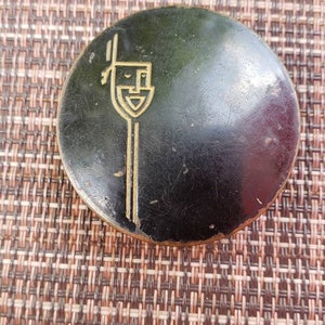 May include: A round, black, compact with a gold design on the top. The design features a stylized image of a building with a tall, thin tower.