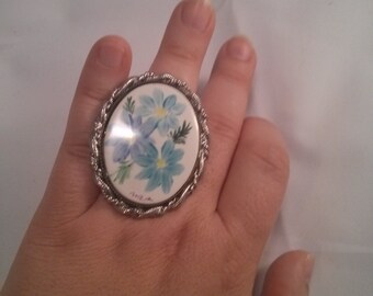 May include: Oval-shaped ring with a floral design. The ring features a silver-colored, twisted rope-like border. The center of the ring has a white background with blue flowers and green leaves. The ring is worn on a hand.