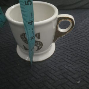 May include: White ceramic mug with a gold handle and trim, featuring a large, ornate letter "S" in a dark color. The mug is approximately 4 inches tall.