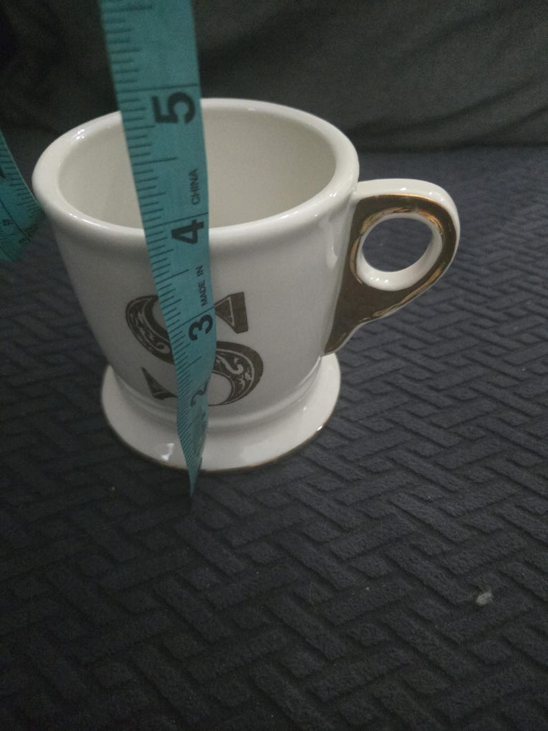 May include: White ceramic mug with a gold handle and trim, featuring a large, ornate letter "S" in a dark color. The mug is approximately 4 inches tall.