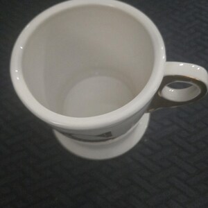 May include: A white ceramic mug with a gold-trimmed handle and base. The mug has a cylindrical shape with a wide opening and a small design near the base. The mug is empty and sits on a dark surface.