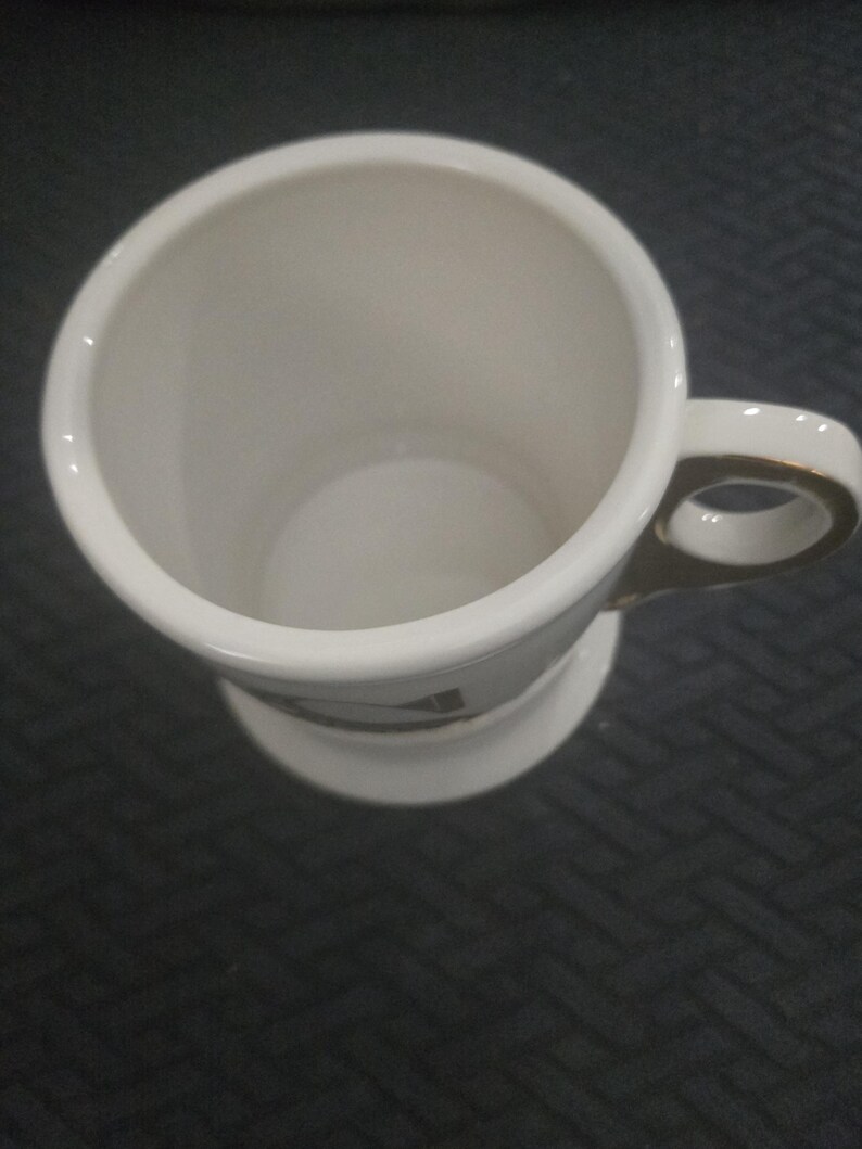 May include: A white ceramic mug with a gold-trimmed handle and base. The mug has a cylindrical shape with a wide opening and a small design near the base. The mug is empty and sits on a dark surface.