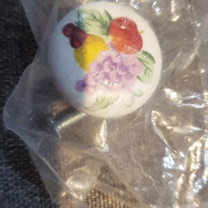 May include: White ceramic cabinet knob with a colorful fruit design. The knob features a red apple, yellow lemon, purple grapes, and green leaves.