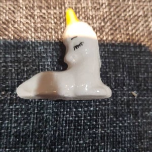 May include: A small white ceramic figurine of a unicorn with a yellow horn and closed eyes. The unicorn is lying down with its head turned to the side.
