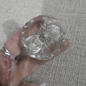 May include: A clear glass cabinet knob with a flower design. The knob has a round shape with a detailed floral pattern and a silver-colored center. The glass is transparent, allowing light to pass through.