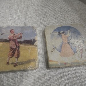 May include: Two square coasters with vintage golf-themed illustrations. One depicts a golfer in a brown suit swinging a club on a green field. The other shows a woman in a dress and hat, also golfing. The coasters have a distressed, aged appearance.