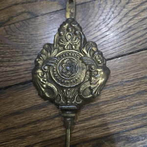 May include: Ornate brass clock pendulum with a decorative floral and scroll design. The pendulum has a loop at the top for hanging and a threaded rod at the bottom. The brass has a warm, aged patina.