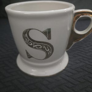 May include: White ceramic mug with a gold handle and rim, featuring a decorative letter "S" in a dark gray color. The mug has a pedestal base and is designed for beverages.