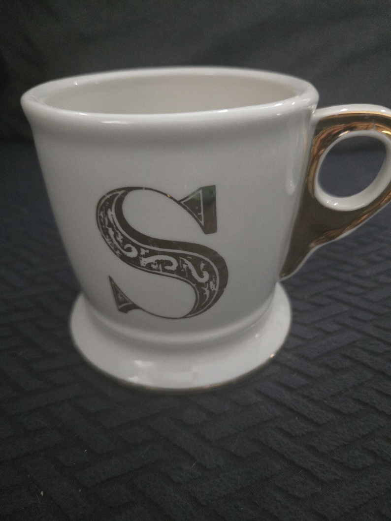 May include: White ceramic mug with a gold handle and rim, featuring a decorative letter "S" in a dark gray color. The mug has a pedestal base and is designed for beverages.
