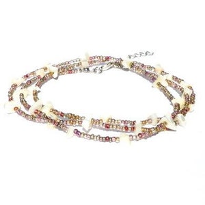 Op de afbeelding: Een meerstrengs ketting met kleine, ronde, metallic kralen in tinten brons, koper en donkerrood, afgewisseld met onregelmatige, gebroken witte, platte, schelpachtige stukjes. De ketting is voorzien van een zilverkleurige metalen karabijnsluiting.