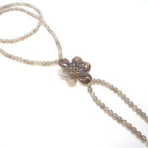 May include: A gray agate necklace with a carved flower pendant. The necklace is made of small, round beads and has a long, tassel-like drop.