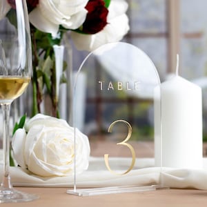 Op de afbeelding: Duidelijke acryl tafelnummerhouder met een gouden 'Tafel 3' inscriptie. De houder staat op een wit tafelkleed met witte kaarsen en een boeket witte en rode rozen.