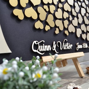 Puede incluir: Un letrero de madera negro con un árbol dorado hecho de corazones. El letrero dice "Quinn & Victor" y "June 16th, 2018".