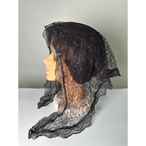 May include: A black lace head covering featuring a delicate floral pattern throughout the fabric. The head covering has a scalloped edge detail along the perimeter and is draped over a mannequin head to display its shape and texture.