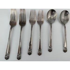 May include: A set of six silver-toned metal utensils comprising four forks and two spoons. Each piece features a slender handle with a decorative floral motif at the base, characterised by a tarnished finish and classic, symmetrical design elements.
