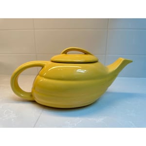 May include: A bright yellow ceramic teapot with a smooth, glossy finish. The teapot features a rounded body with horizontal ribbed detailing, a curved handle on the left side, a pointed spout on the right, and a matching lid with a small handle.